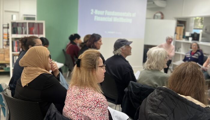 Mixed group of adults sit in room listening to Workshop presenter, with a presentation screen visible in the background.